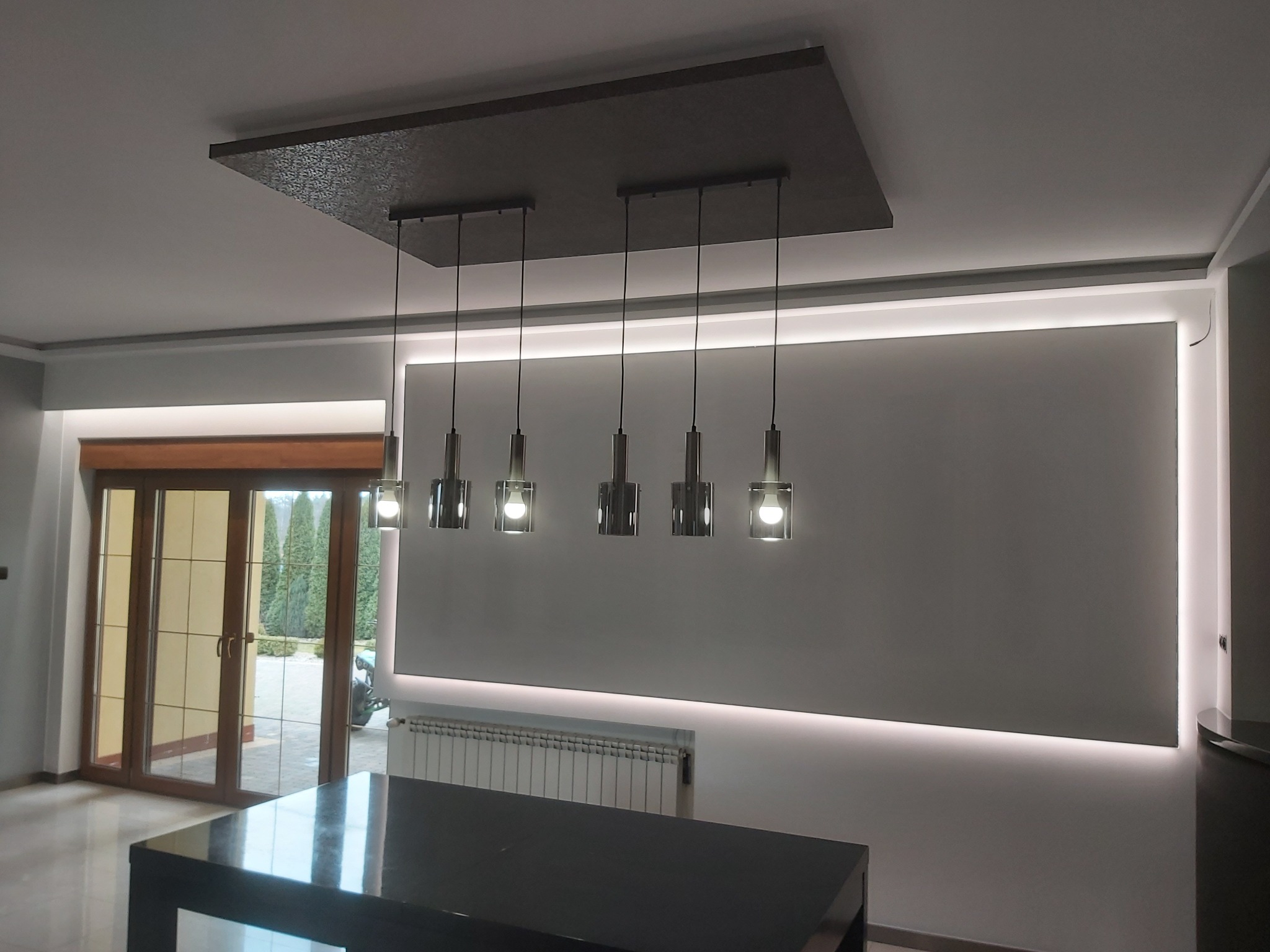 Nowoczesne oświetlenie sufitowe z pięcioma lampami wiszącymi, podświetlana ściana i drzwi balkonowe z widokiem na zewnątrz.