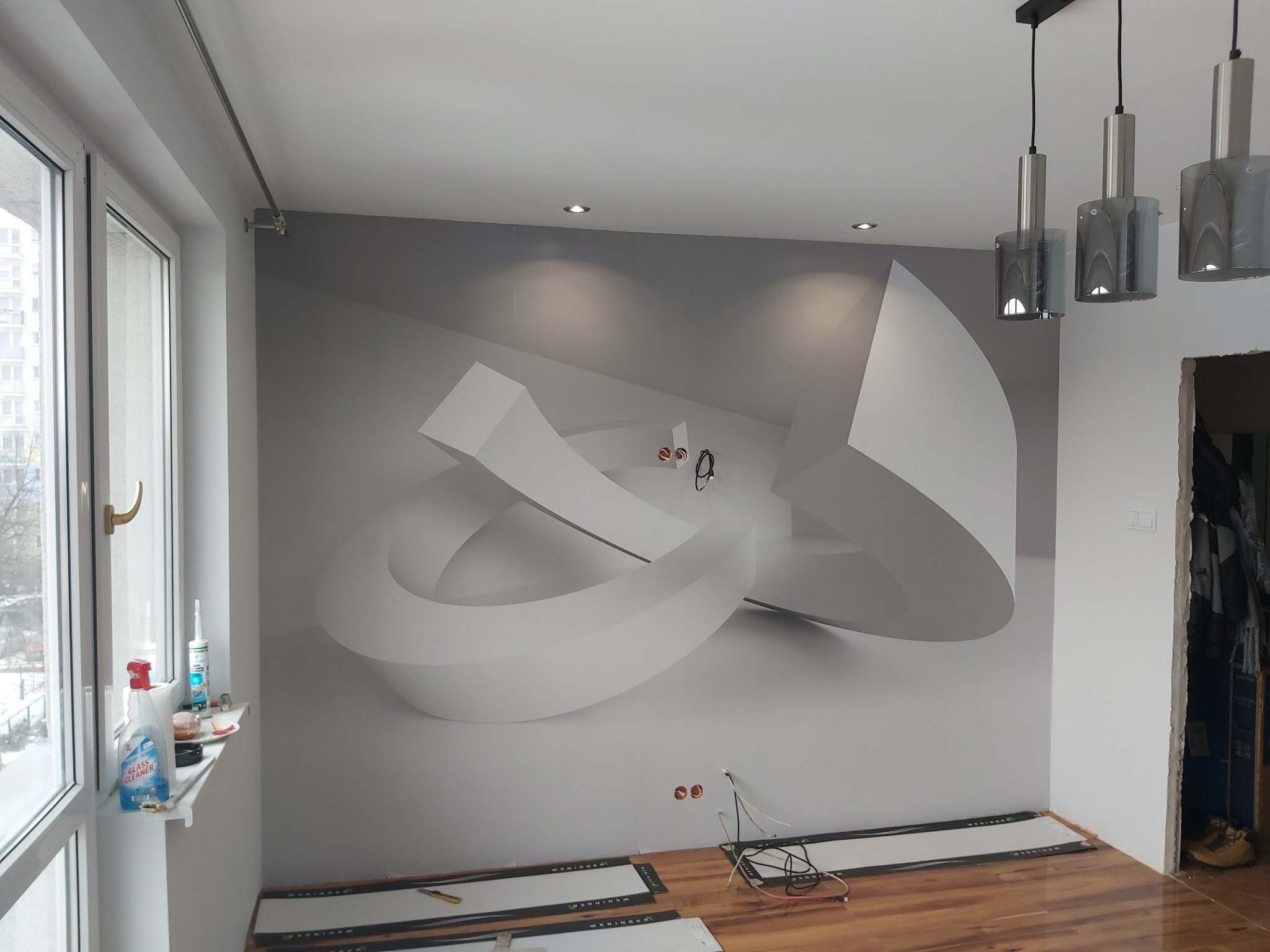 Pokój w trakcie prac wykończeniowych: panele podłogowe częściowo ułożone, na ścianie fototapeta z motywem abstrakcyjnym, widoczne narzędzia i chemia gospodarcza na parapecie okna.