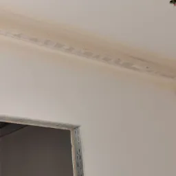 Niedokończone prace wykończeniowe: widoczna konstrukcja sufitu podwieszanego z profilami i częściowo nałożoną masą szpachlową, surowy oścież metalowy drzwiowy, czujnik ruchu i detektor dymu...