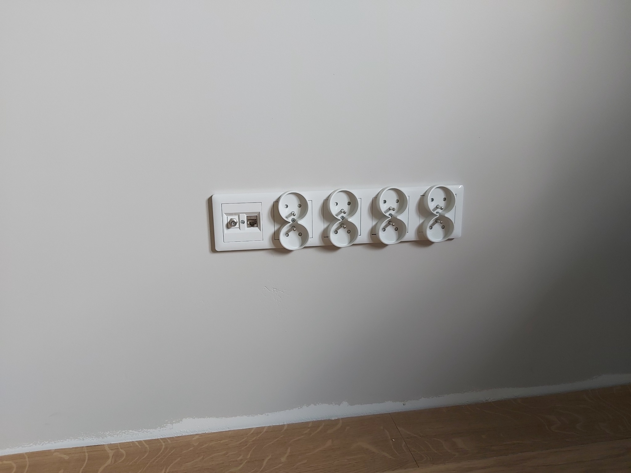 Biała listwa zasilająca z czterema gniazdami elektrycznymi i gniazdem antenowym/internetowym, zamontowana na jasnej ścianie nad drewnianą podłogą.