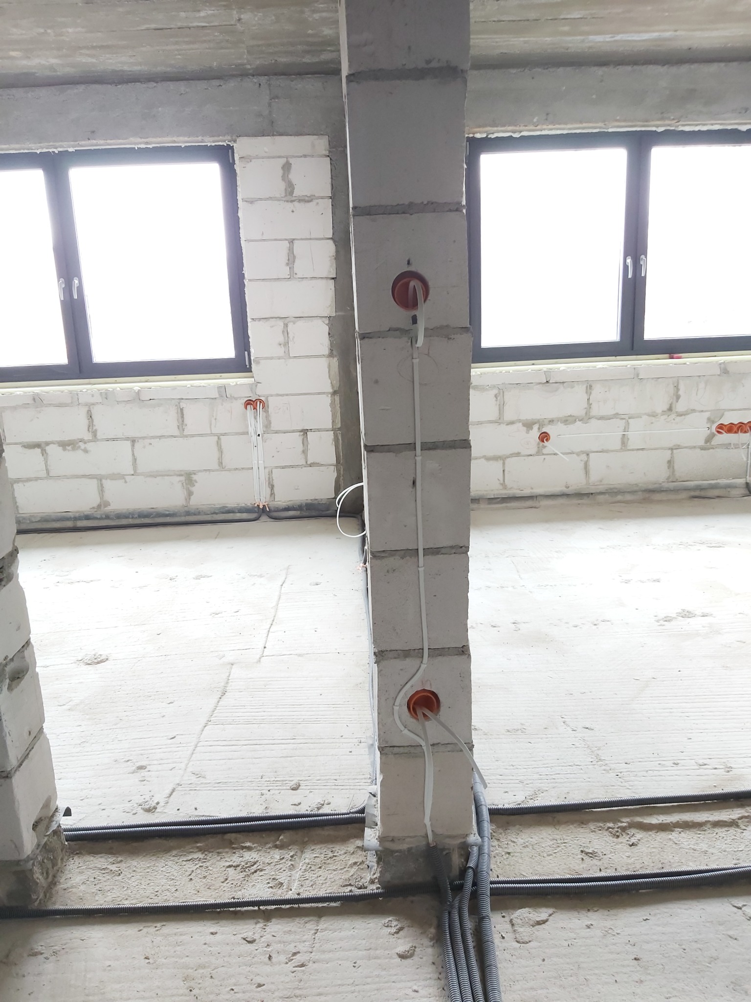 Surowy stan wnętrza w trakcie instalacji elektrycznej: widoczne kable w peszlach, puszki instalacyjne w ścianach z betonu komórkowego, okna z czarnymi ramami.