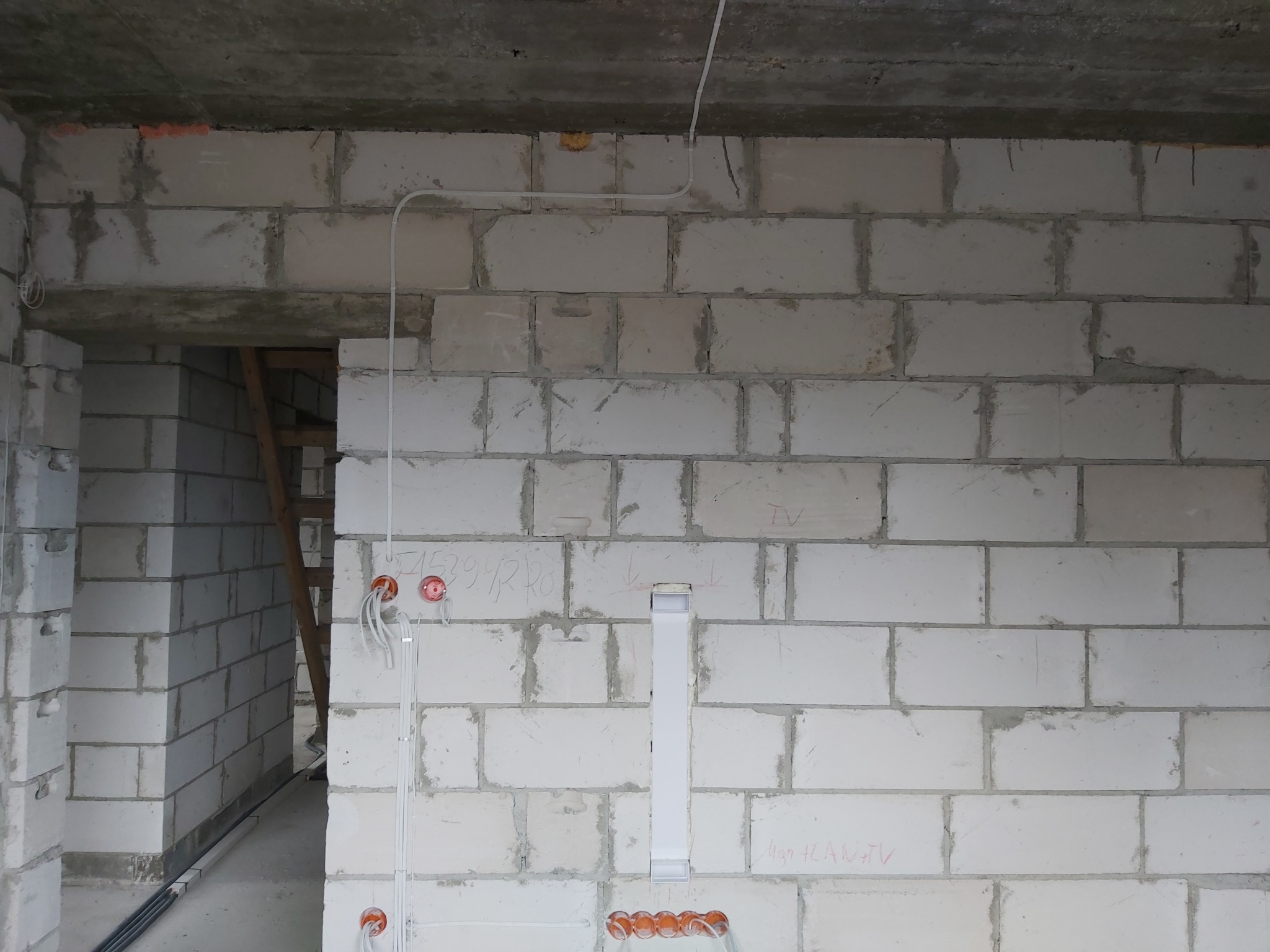 Surowy mur z betonu komórkowego w trakcie prac instalacyjnych: widoczne puszki podtynkowe z przewodami, peszel z kablami na podłodze i rurka instalacyjna na ścianie, sufit betonowy.