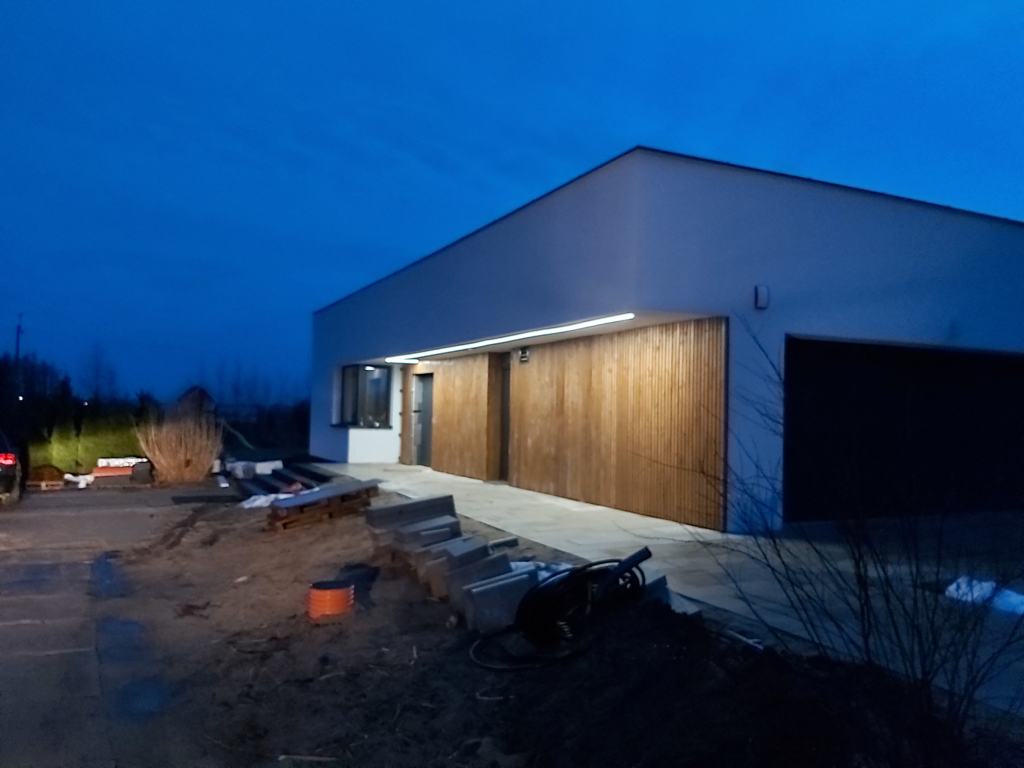 Nowoczesny dom jednorodzinny z drewnianą elewacją i oświetleniem zewnętrznym, widoczny wczesnym wieczorem, teren wokół domu w trakcie prac wykończeniowych.