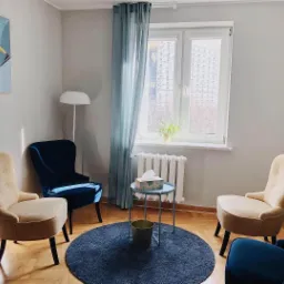 Jasne wnętrze gabinetu z czterema fotelami (dwa beżowe, dwa granatowe) ustawionymi wokół okrągłego, niebieskiego dywanu. Na dywanie mały, okrągły stolik z chusteczkami. Na ścianie abstrakcyjny obraz.