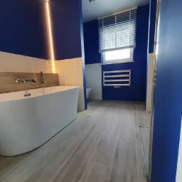 Nowoczesna łazienka z wolnostojącą białą wanną, niebieskimi ścianami z oświetleniem LED, płytkami imitującymi drewno na podłodze i grzejnikiem drabinkowym pod oknem z żaluzjami.