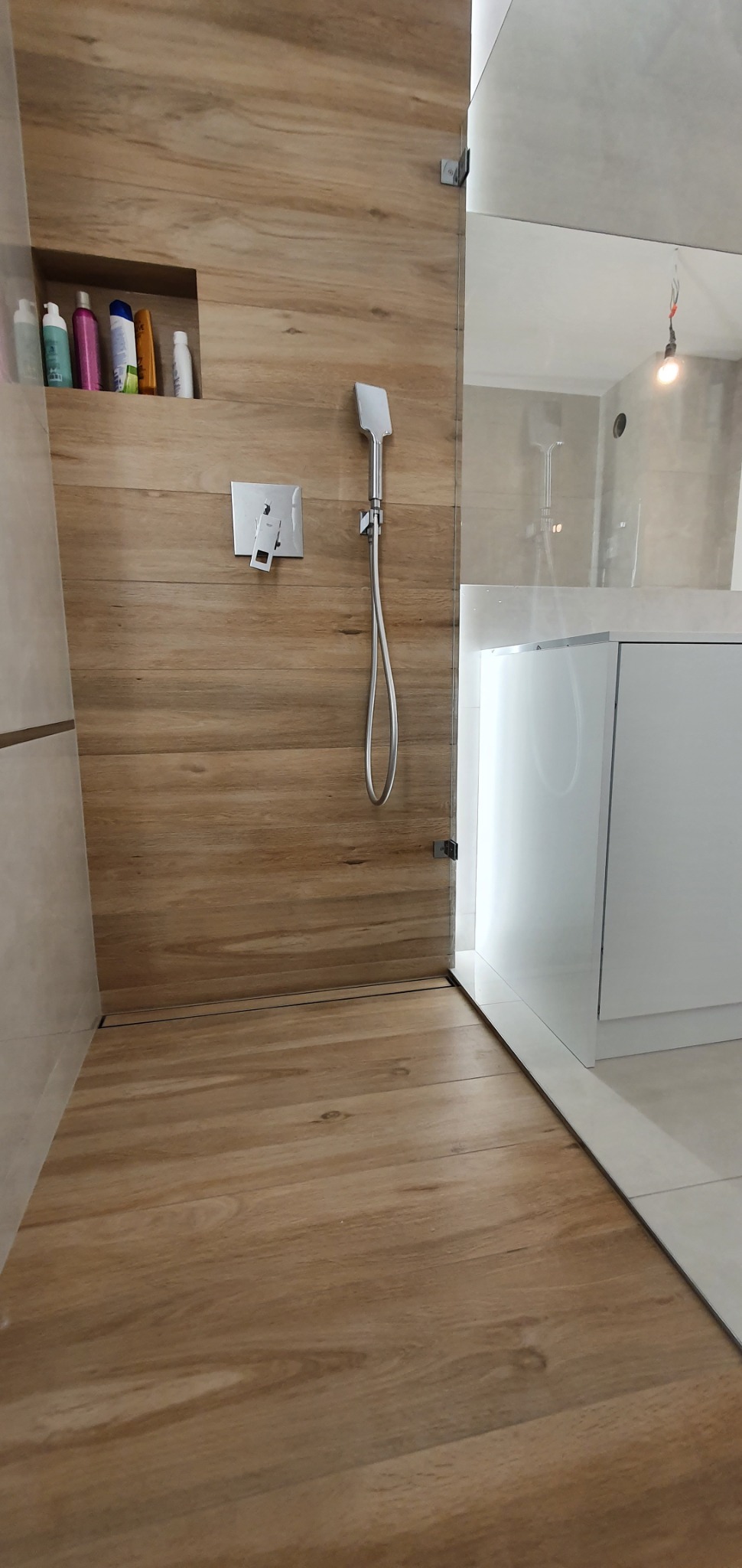 Nowoczesna kabina prysznicowa walk-in z odpływem liniowym w podłodze wyłożonej płytkami imitującymi drewno, bateria podtynkowa i deszczownica na ścianie z wnęką na kosmetyki.