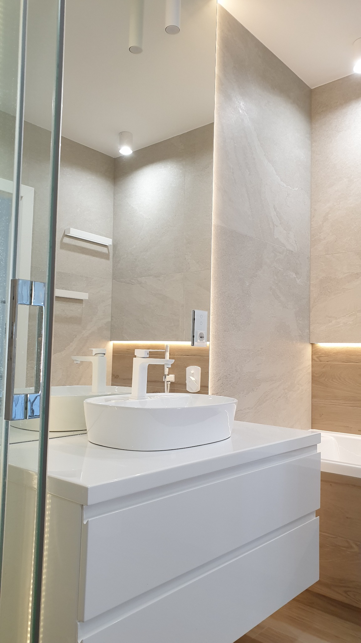 Nowoczesna łazienka z owalną umywalką nablatową na białej szafce z szufladami, kremowe płytki na ścianach, oświetlenie LED pod lustrem i półkami, widoczny fragment kabiny prysznicowej.