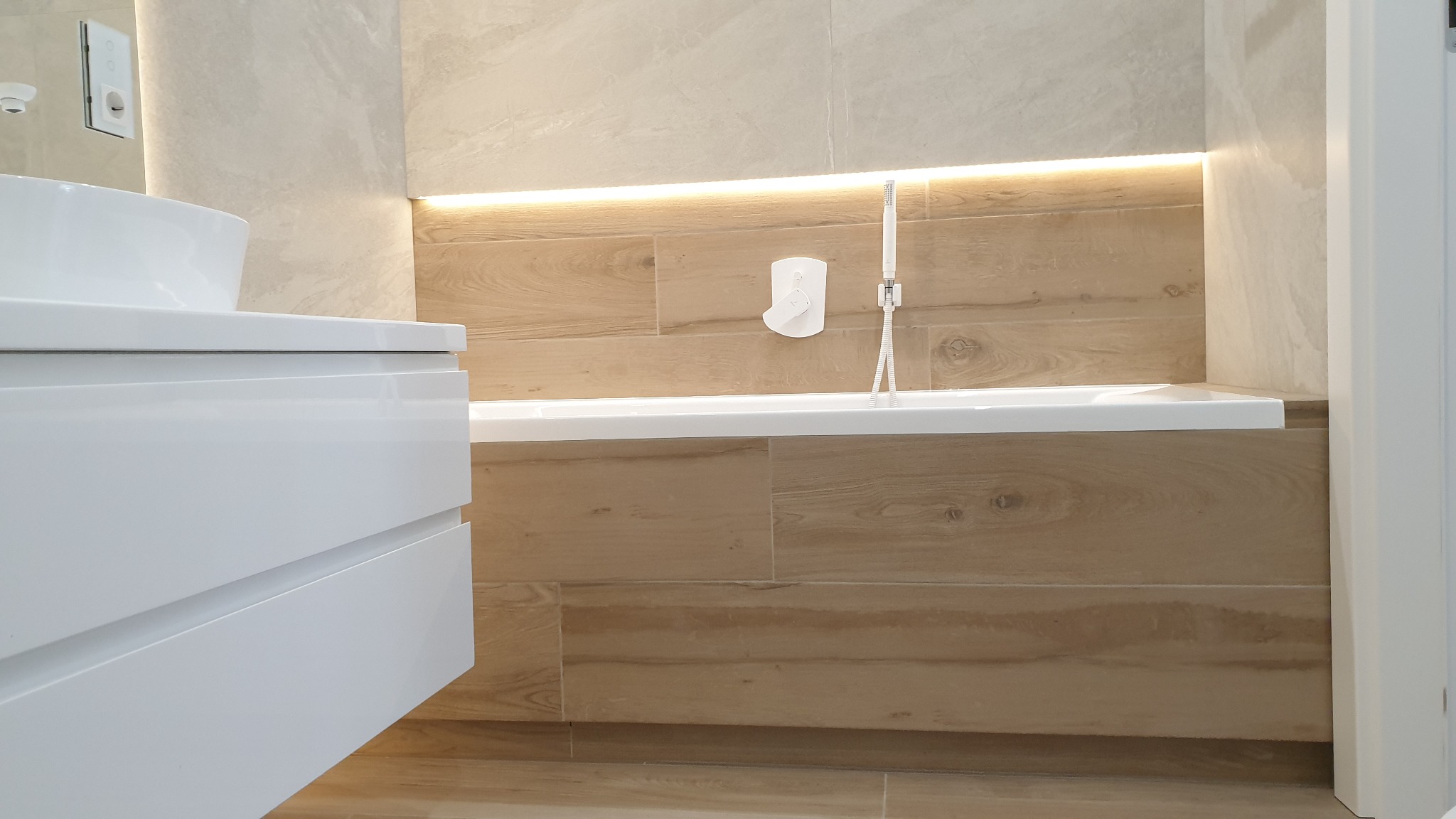 Nowoczesna łazienka z wanną obudowaną płytkami imitującymi drewno, podświetlona listwą LED, z białą umywalką na szafce i armaturą.