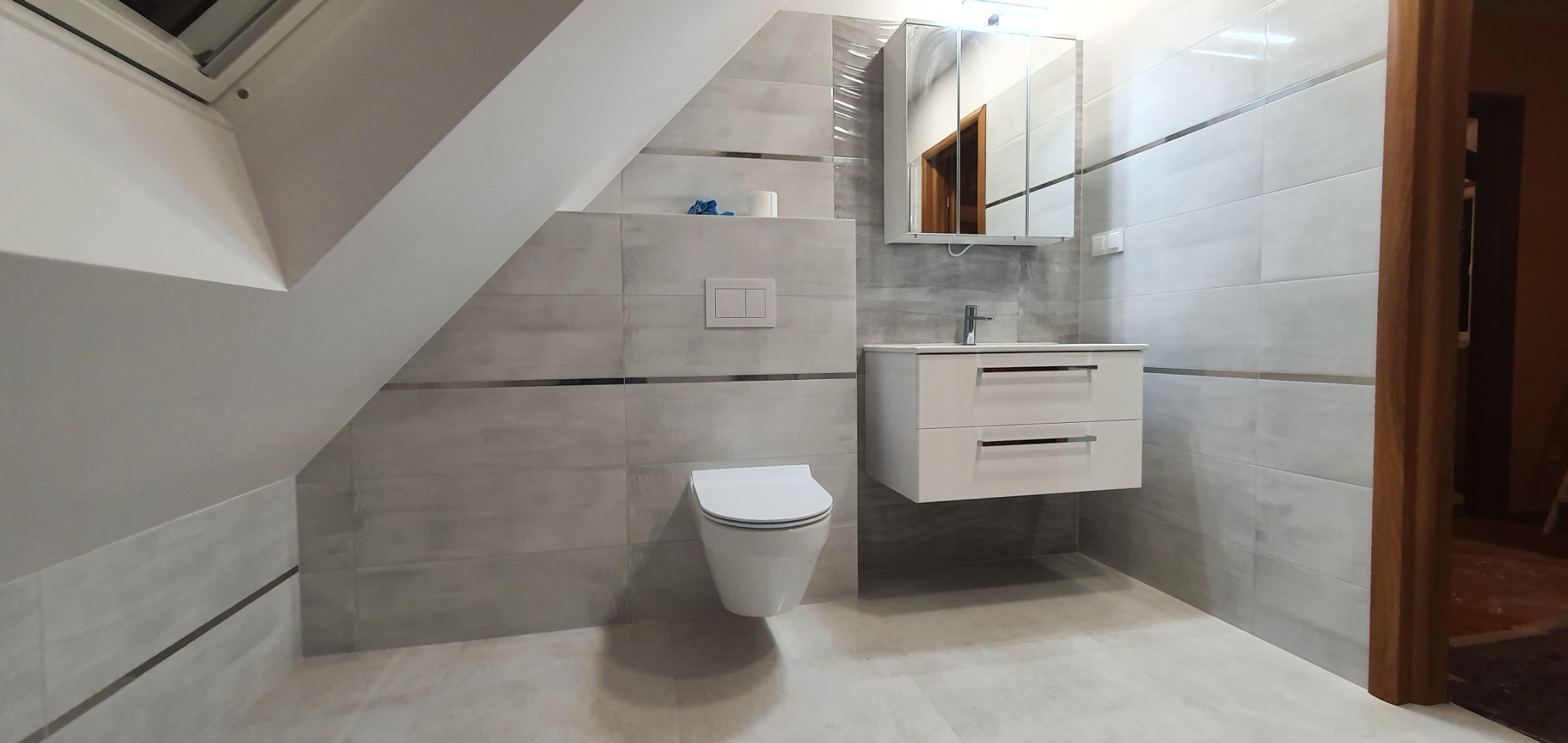 Wykończona łazienka na poddaszu ze skośnym sufitem, szare płytki z poziomymi metalowymi listwami, podwieszana toaleta, biała szafka z umywalką i lustrem, widok na otwarte drzwi.