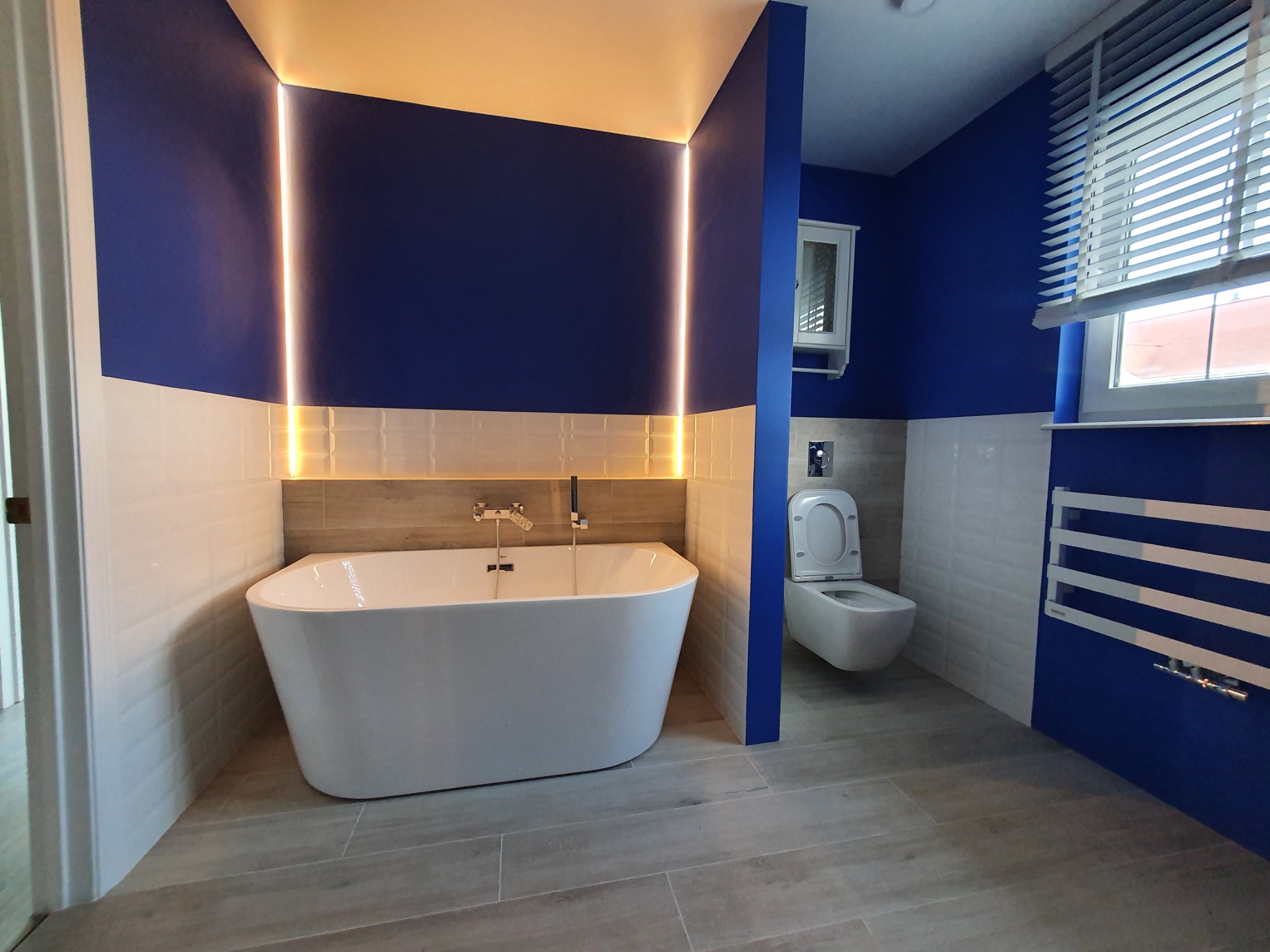 Nowoczesna łazienka z wolnostojącą wanną, podświetleniem LED na ścianie w kolorze głębokiego granatu, jasnymi płytkami imitującymi drewno na podłodze i ścianach, oraz toaletą w tle.