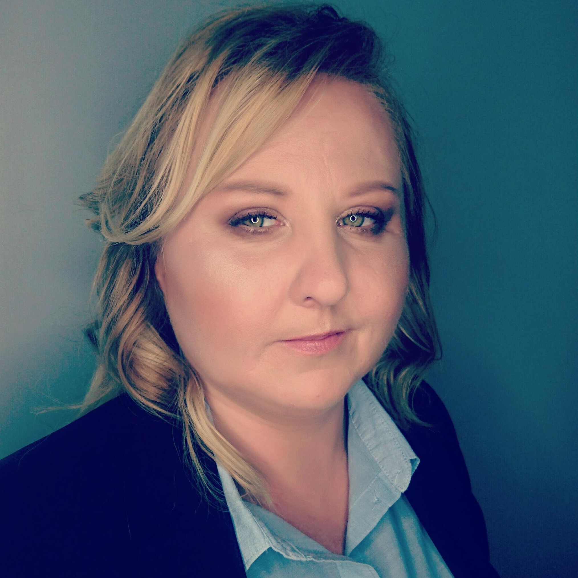 Portret kobiety z blond włosami, ubranej w błękitną koszulę i czarną marynarkę, na jednolitym tle.