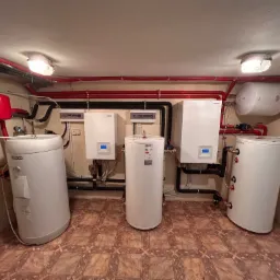 Kotłownia z trzema zbiornikami akumulacyjnymi ciepła, dwoma piecami i instalacją rurową w kolorach czerwonym i czarnym, oświetlona dwoma lampami sufitowymi.