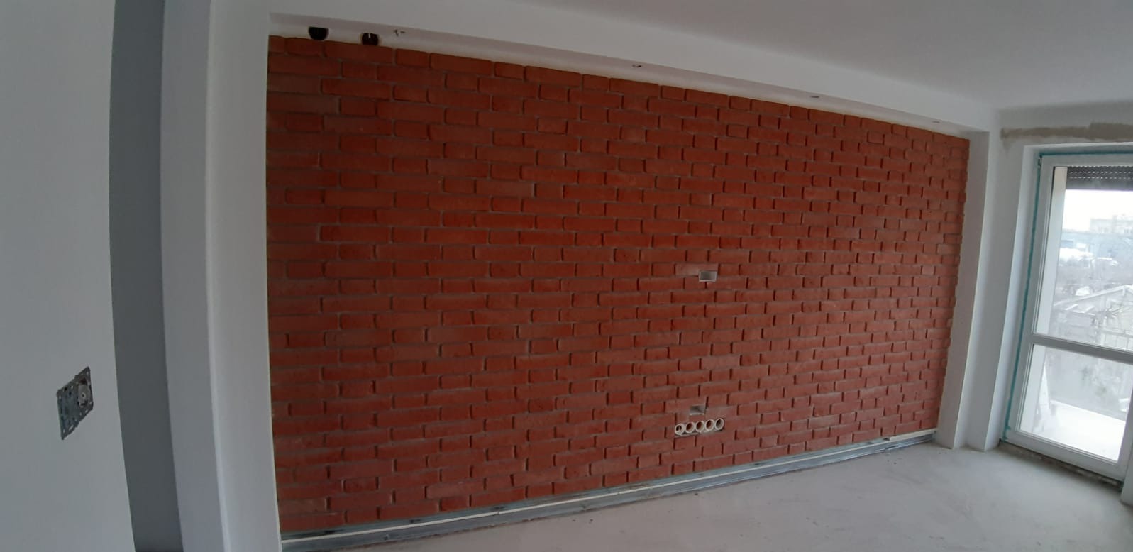 Ściana wyłożona czerwoną cegłą dekoracyjną w nowym pomieszczeniu, widoczne puszki elektryczne i otwory pod instalacje, surowy stan wykończenia.
