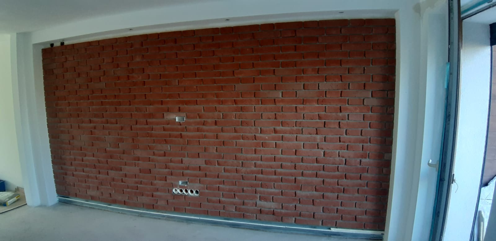 Ściana z czerwonej cegły z widocznymi otworami na gniazdka elektryczne, w trakcie prac wykończeniowych, widoczne profile podłogowe.