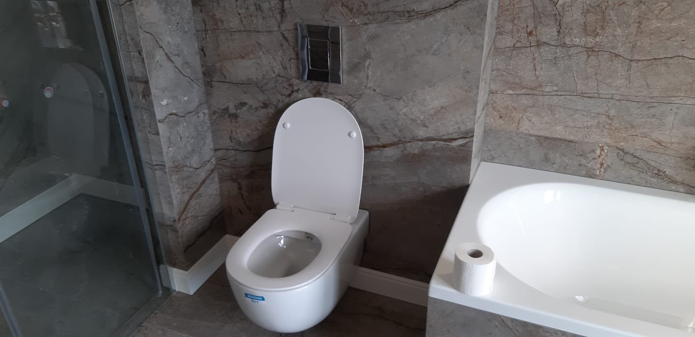 Nowoczesna łazienka z marmurowymi płytkami, wisząca toaleta z otwartą deską, kabina prysznicowa ze szklanymi drzwiami i biała wanna z rolką papieru toaletowego.