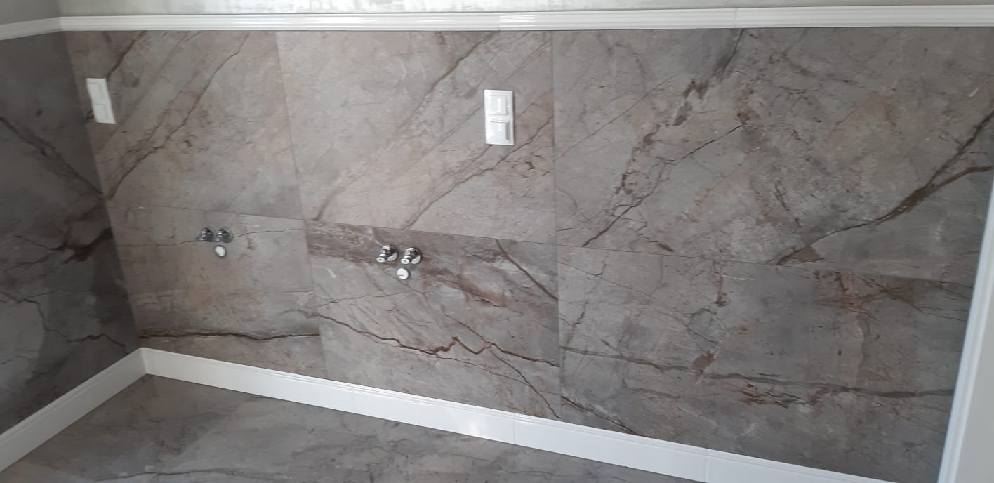 Ściana i podłoga wyłożone dużymi, szarymi płytkami imitującymi marmur, z widocznymi chromowanymi przyłączami wodnymi i białymi listwami przypodłogowymi oraz gniazdkiem elektrycznym.