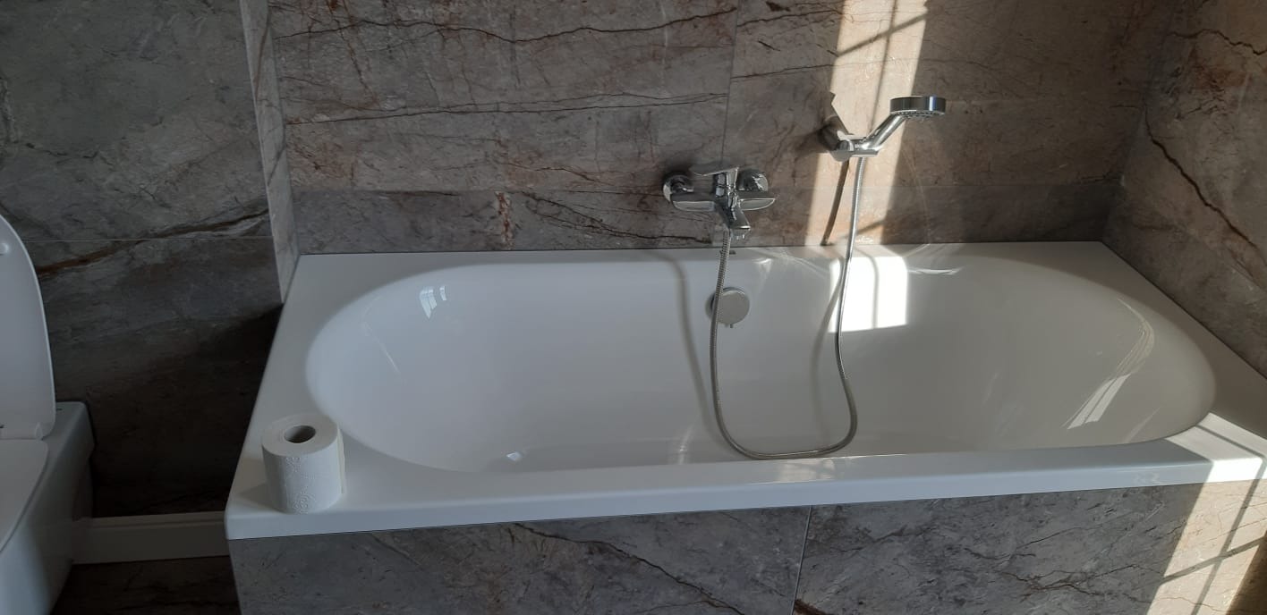 Biała wanna z zamontowaną baterią i słuchawką prysznicową, obok toaleta i rolka papieru toaletowego, ściany wykończone płytkami imitującymi marmur w odcieniach szarości i beżu, oświetlone...