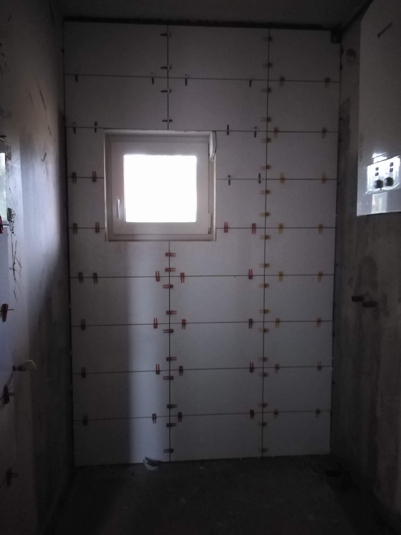 Ściana łazienki w trakcie układania białych płytek z użyciem krzyżyków dystansowych, widoczne okno i surowy stan ścian w tle.