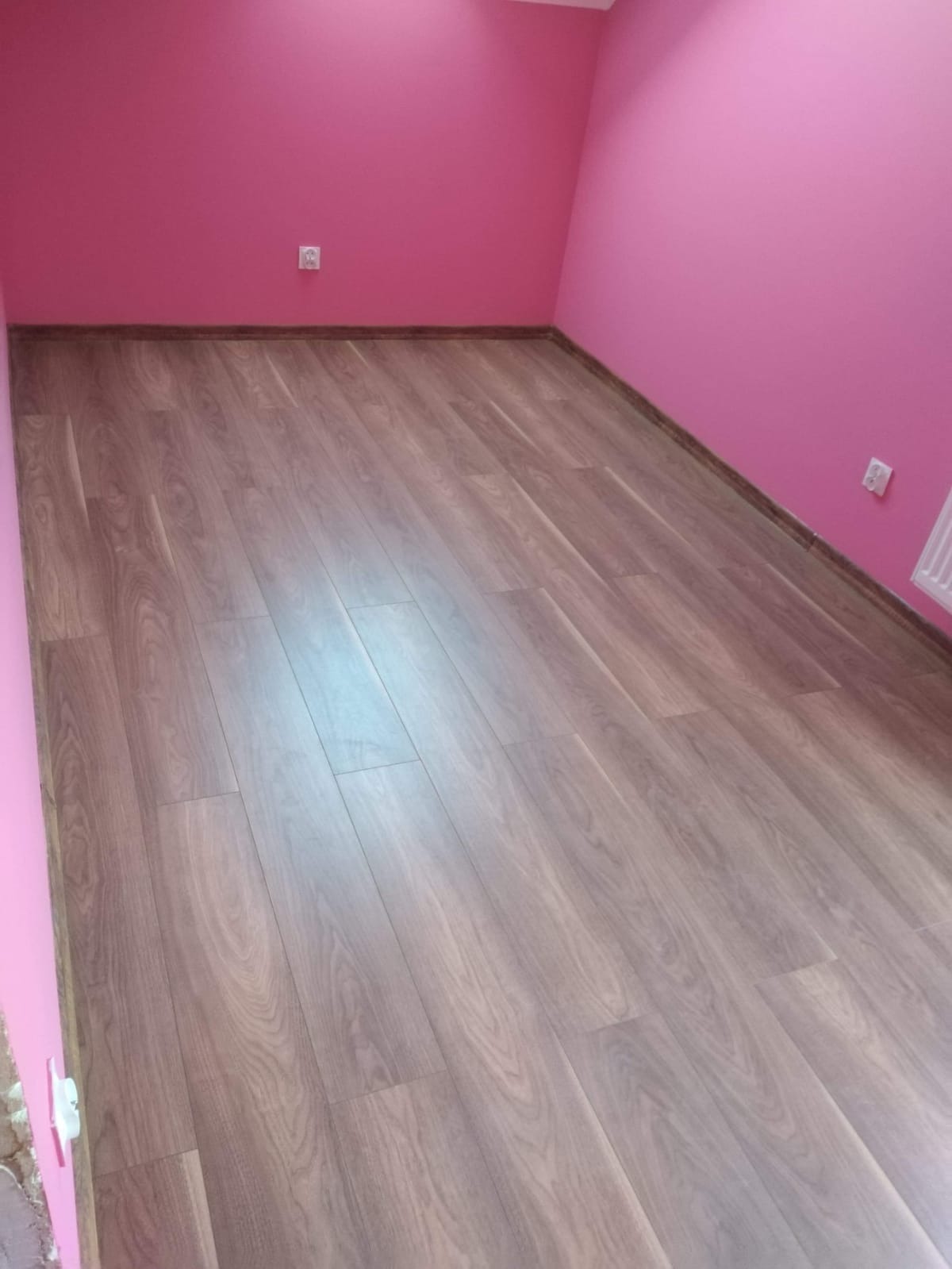Pokój z nowo ułożonymi panelami podłogowymi w odcieniach brązu, ściany pomalowane na intensywny różowy kolor, widoczne gniazdko elektryczne i fragment grzejnika.