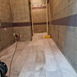 Wnętrze łazienki w trakcie remontu: położone panele podłogowe imitujące drewno, ściany z szarymi płytkami z bordowym pasem, widoczne instalacje hydrauliczne i ż&oacute;łta kuweta malarska.
