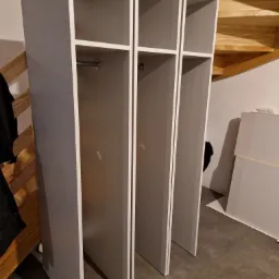 Trzy moduły szafy wnękowej w kolorze szarym, z drążkami na ubrania, ustawione pod drewnianymi schodami w trakcie montażu, widoczne płyty meblowe w tle.