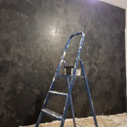 Ściana z dekoracyjną, ciemną fakturą, widoczna drabina malarska z puszką farby, fragment zabezpieczonej podłogi.