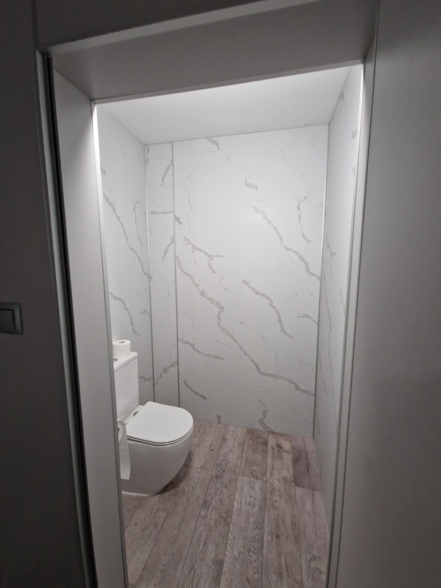 Nowoczesna, mała toaleta z białymi panelami ściennymi imitującymi marmur, podłogą z paneli imitujących drewno i minimalistyczną białą muszlą klozetową, widziana zza krawędzi wejścia.