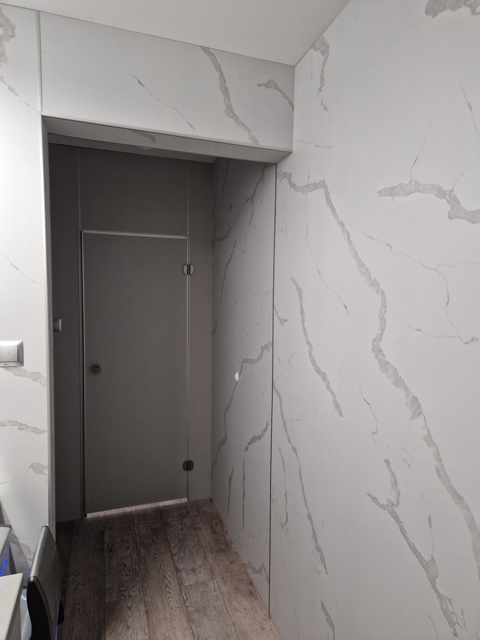 Nowoczesna łazienka z szarą, szklaną kabiną prysznicową, wykończona płytkami imitującymi biały marmur z szarymi żyłami i drewnianą podłogą.