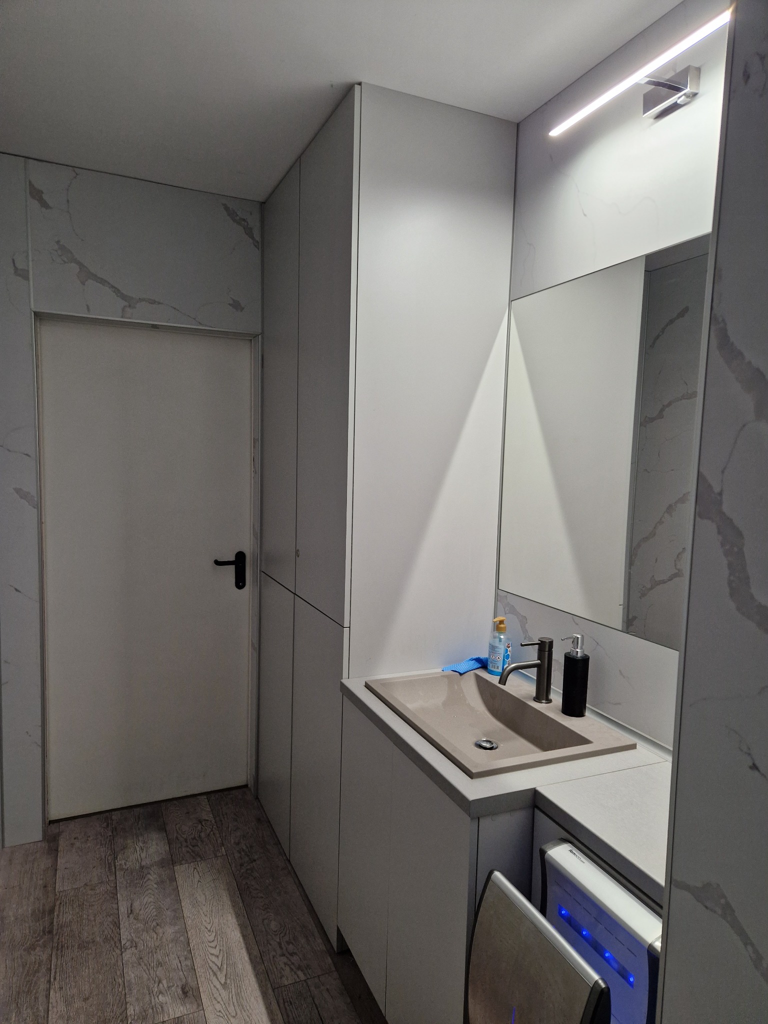 Nowoczesna łazienka z beżową umywalką, lustrem z oświetleniem LED, szarą szafką i pralką pod blatem, wykończona płytkami imitującymi marmur.