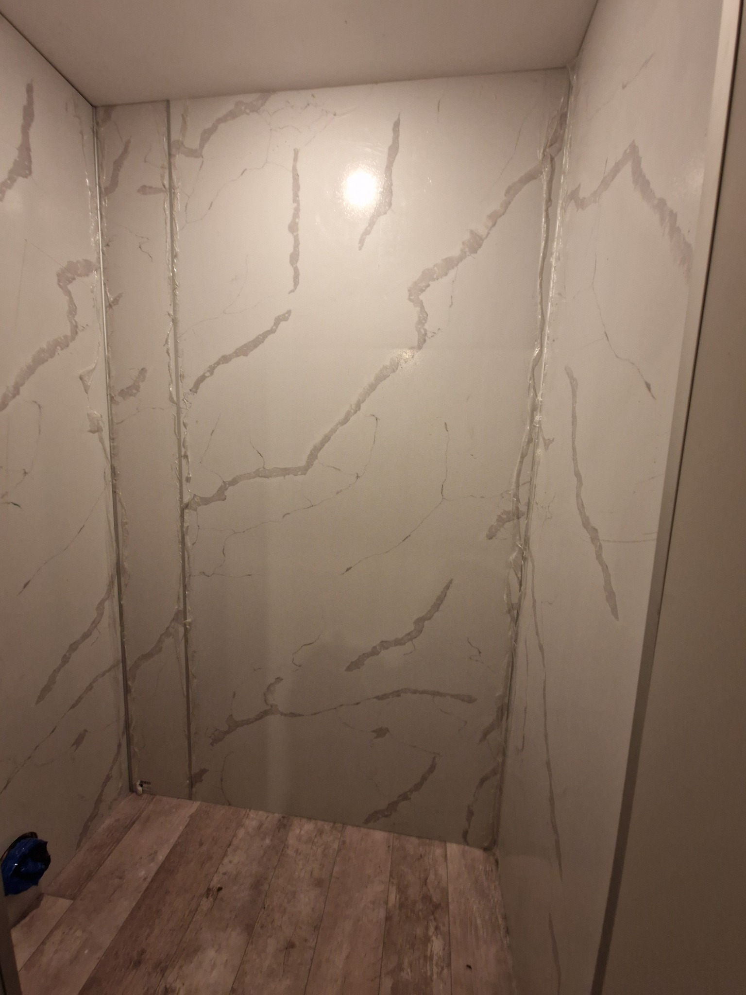 Wykończona kabina prysznicowa z panelami ściennymi imitującymi marmur i drewnopodobną podłogą, widoczne łączenia paneli.