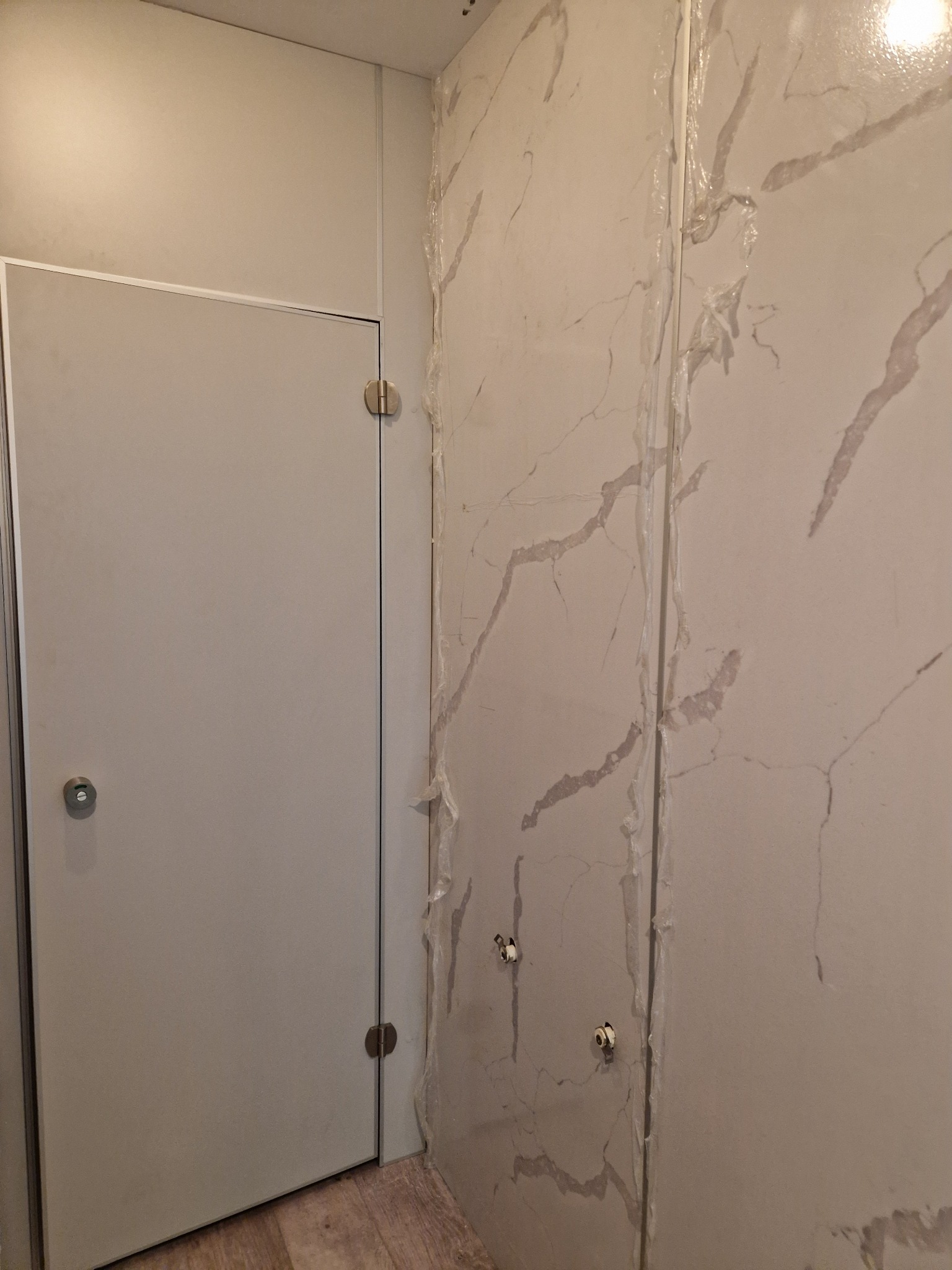 Montaż paneli ściennych imitujących marmur w łazience, z widocznymi punktami mocowania i folią ochronną na panelach, obok zamontowane drzwi.