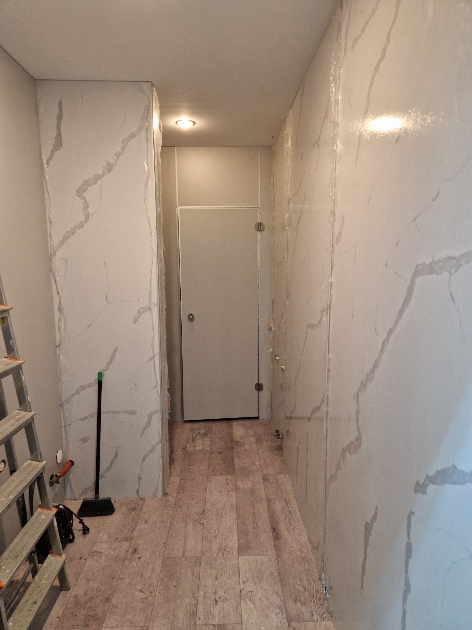 Montaż paneli ściennych z efektem marmuru w wąskim korytarzu, widoczne zabezpieczenia folią, drabina malarska i szczotka na podłodze z imitacją drewna.
