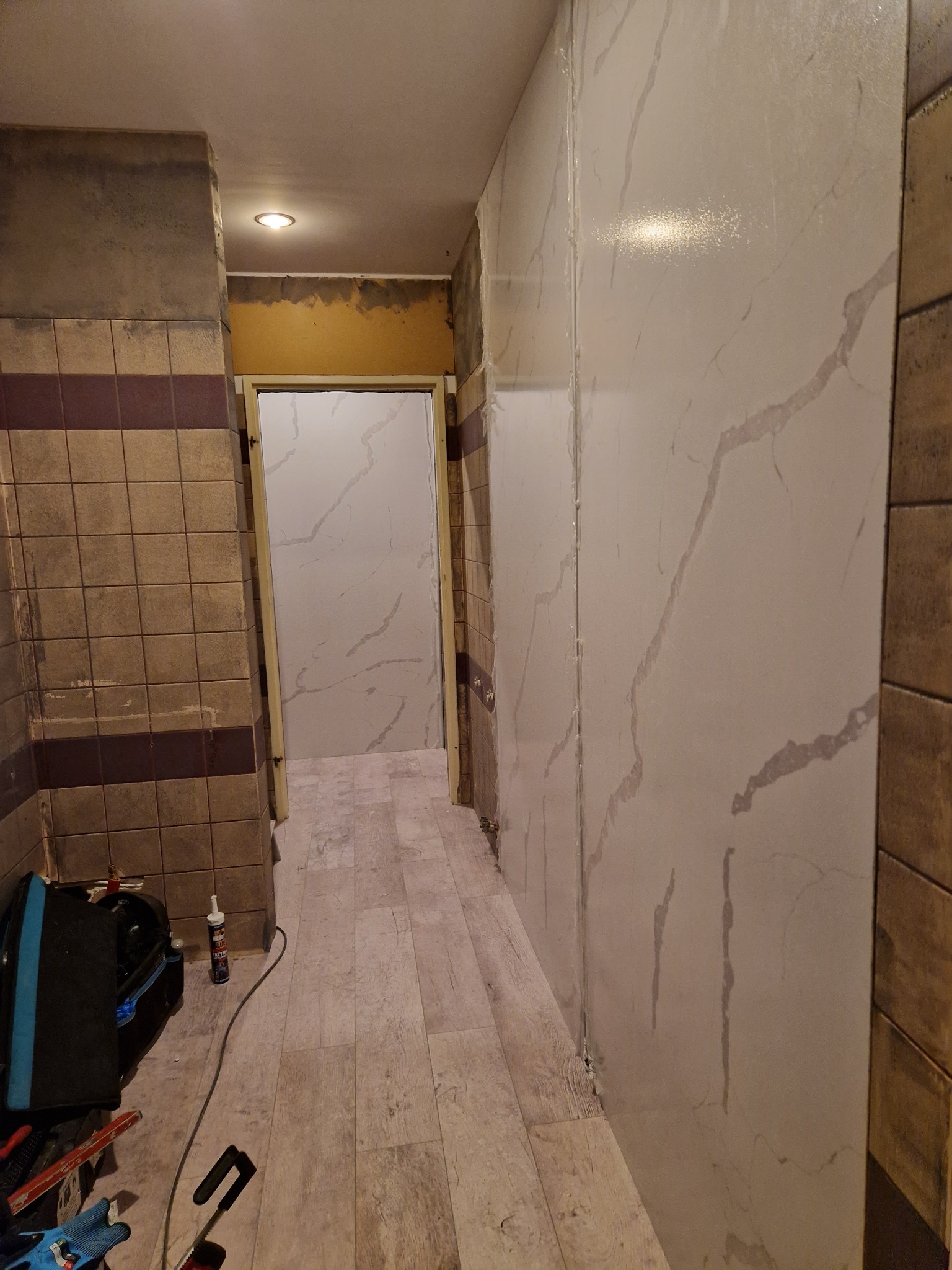 Korytarz w trakcie remontu: nowe panele ścienne imitujące marmur zamontowane częściowo, widoczne narzędzia i materiały budowlane na podłodze, stare płytki na ścianie po lewej stronie.