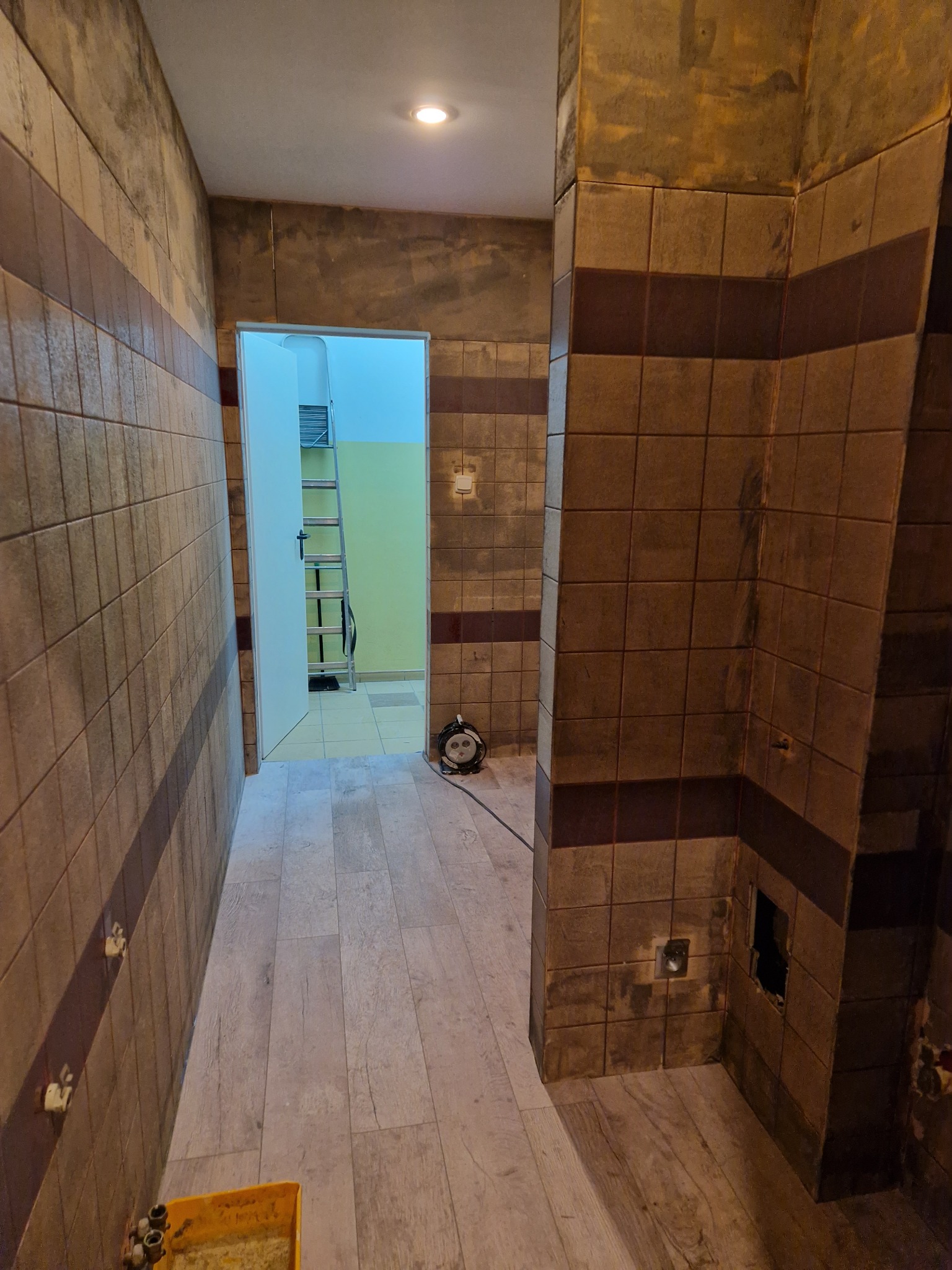 Wnętrze łazienki w trakcie remontu, widoczne brązowe płytki z ciemniejszym pasem, drewnopodobna podłoga, drabina w przejściu do innego pomieszczenia, ściany częściowo przygotowane do malowania...