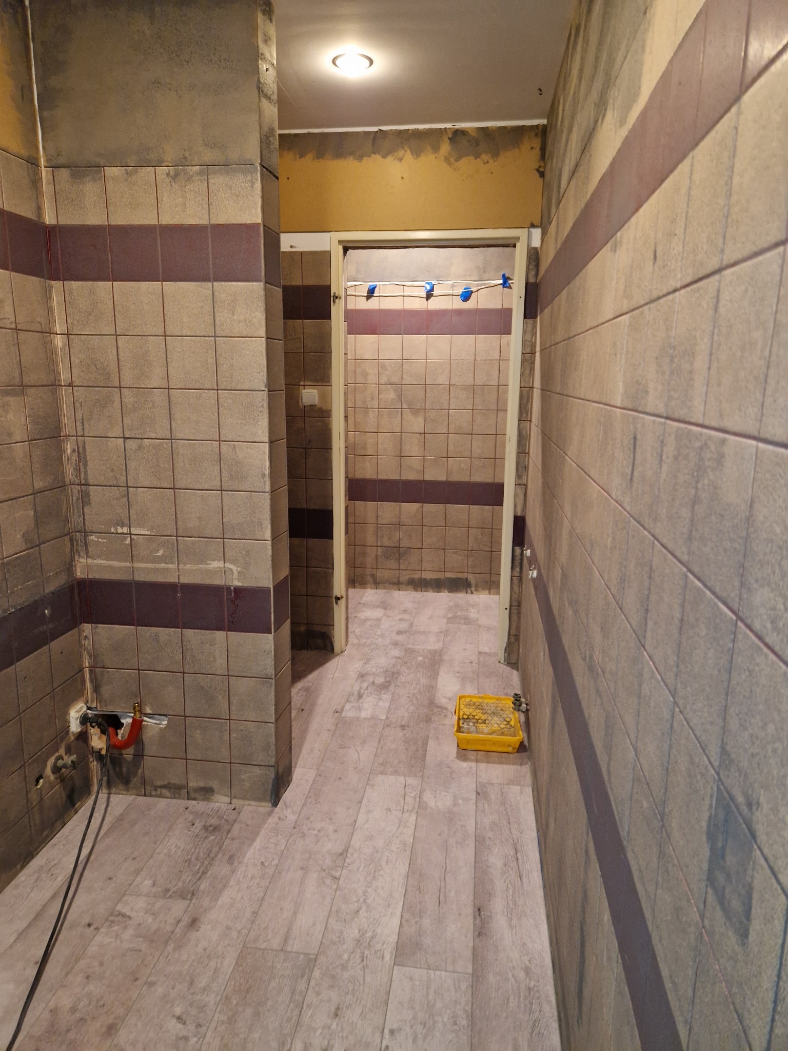 Wnętrze łazienki w trakcie remontu; widoczne płytki na ścianach z ciemnym pasem, podłoga imitująca drewno, żółta kuweta malarska i wystające rury. Na ścianach widoczne ślady po remoncie.