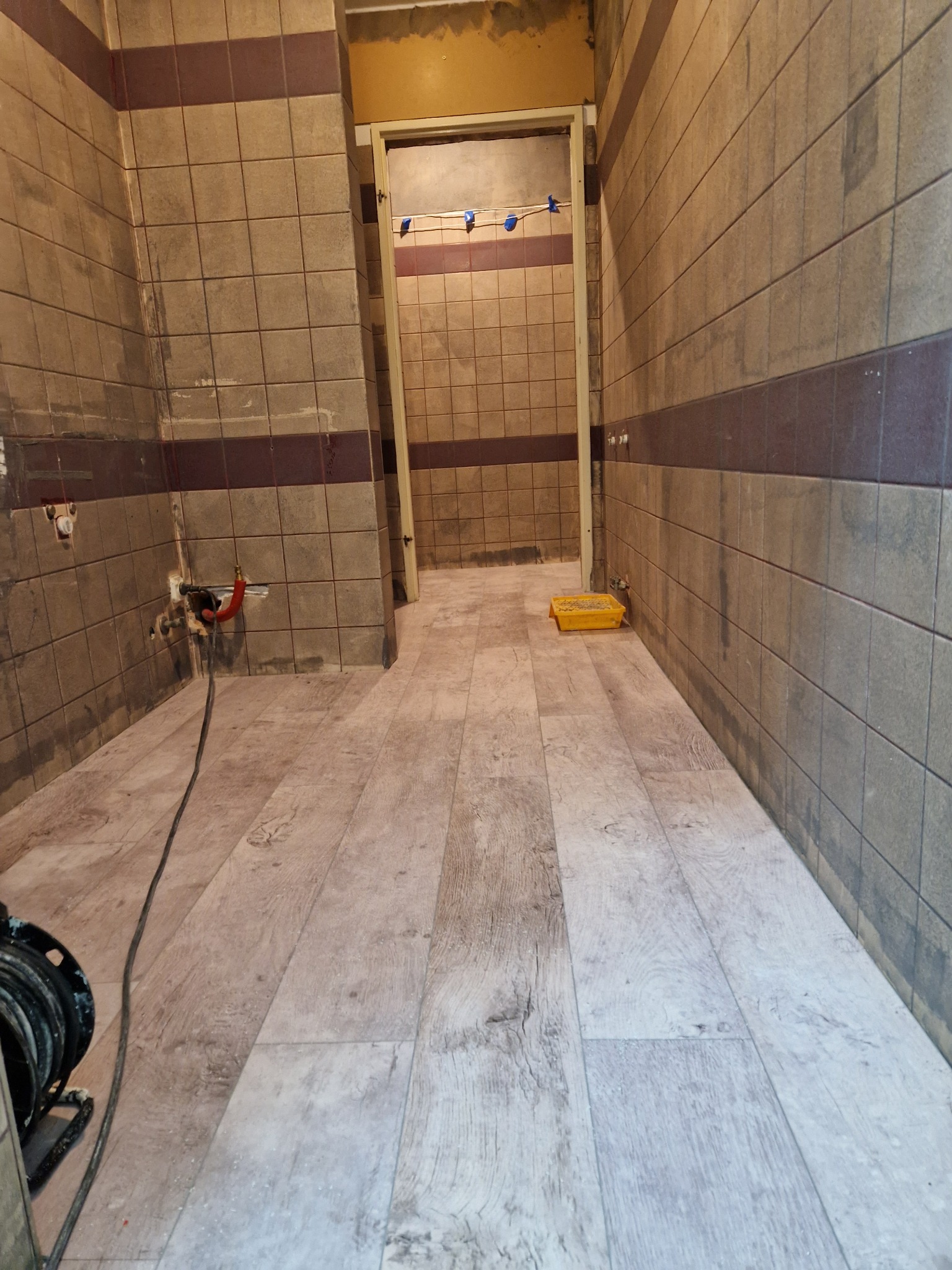 Wnętrze łazienki w trakcie remontu: położone panele podłogowe imitujące drewno, ściany z szarymi płytkami z bordowym pasem, widoczne instalacje hydrauliczne i żółta kuweta malarska.