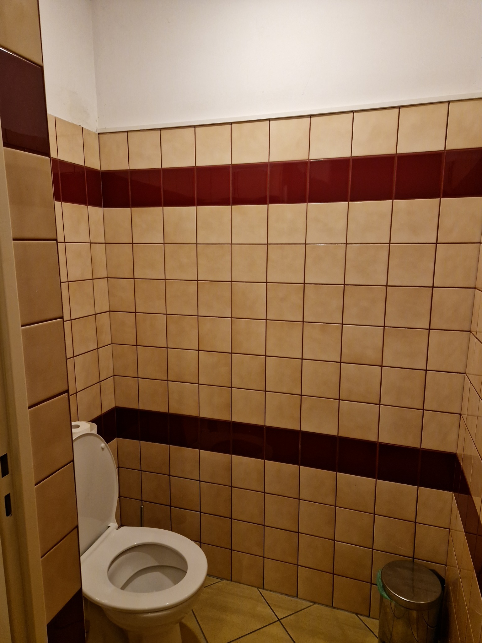 Wykończona łazienka z beżowymi płytkami i bordowym pasem, widoczna toaleta i kosz na śmieci.