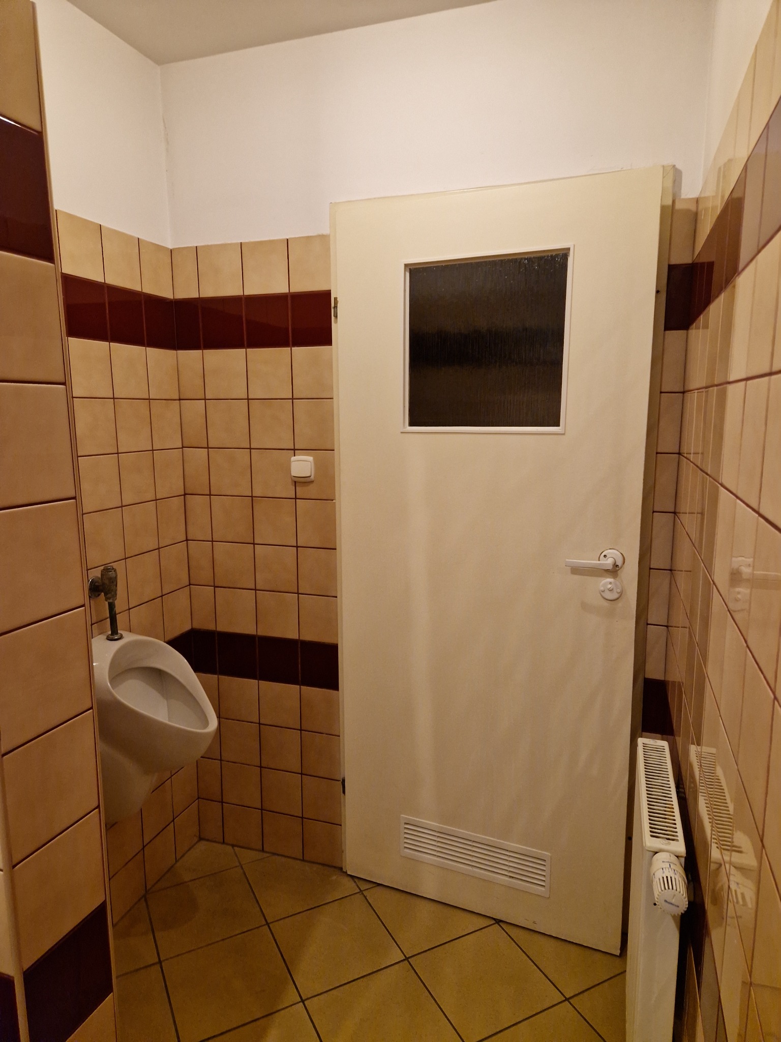 Ukończona toaleta publiczna z beżowymi kafelkami z brązowym paskiem, białym pisuarzem, drzwiami z małym okienkiem i grzejnikiem żeberkowym.