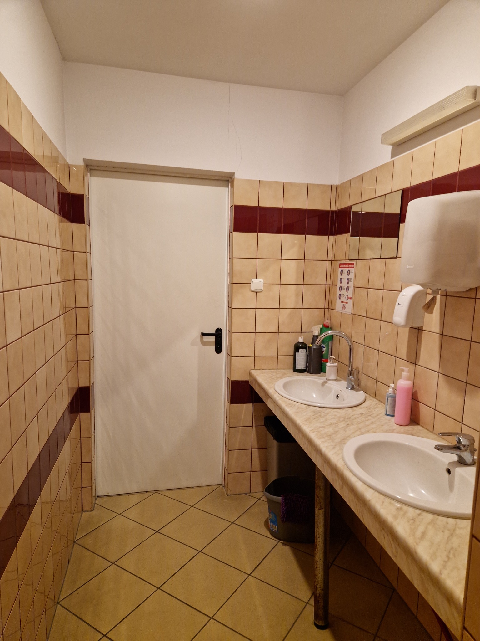 Umywalki w publicznej toalecie wyłożonej beżowymi płytkami z bordowymi pasami, widoczna biała ściana z drzwiami i dozownik do papieru.