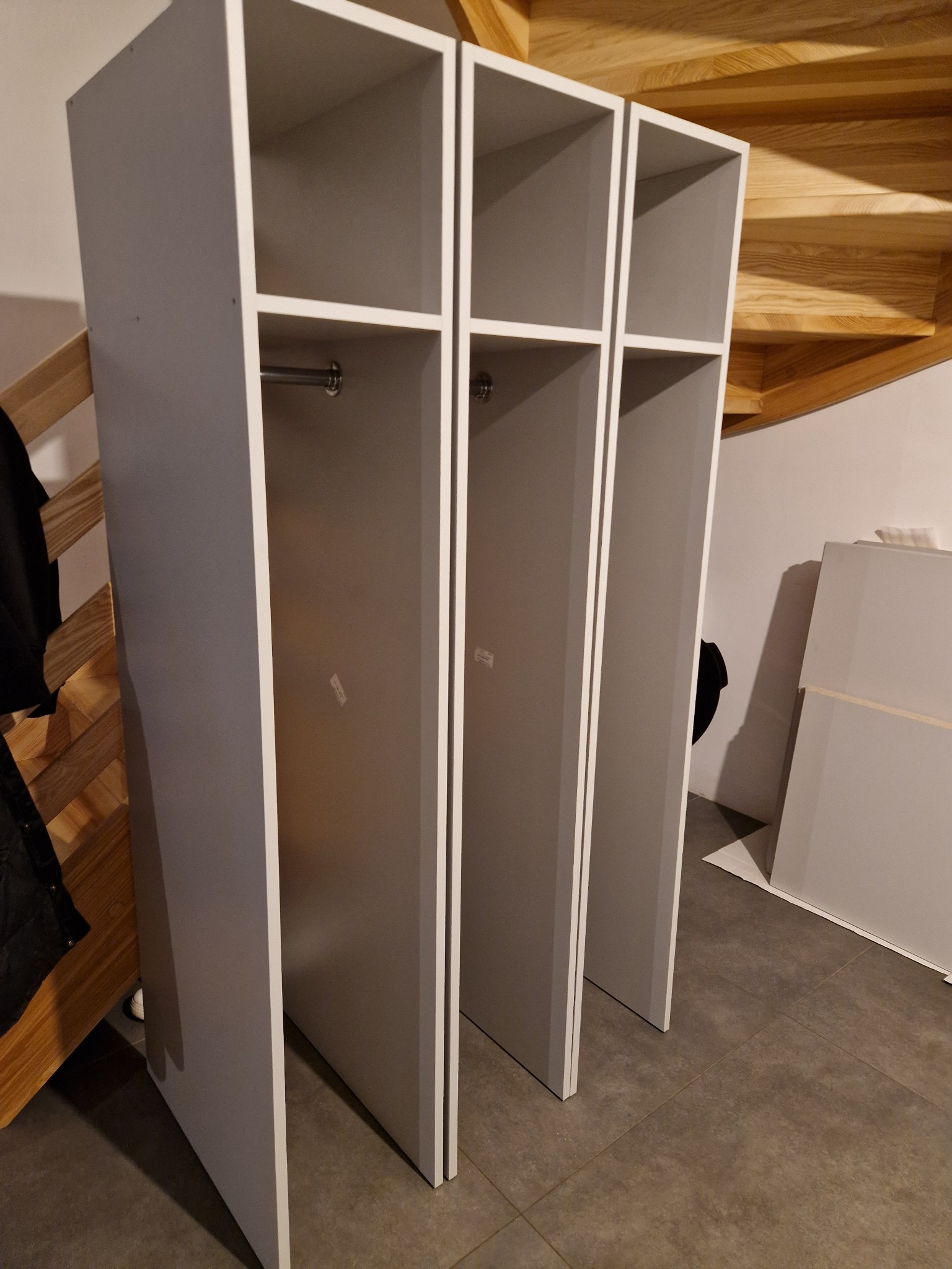 Trzy moduły szafy wnękowej w kolorze szarym, z drążkami na ubrania, ustawione pod drewnianymi schodami w trakcie montażu, widoczne płyty meblowe w tle.
