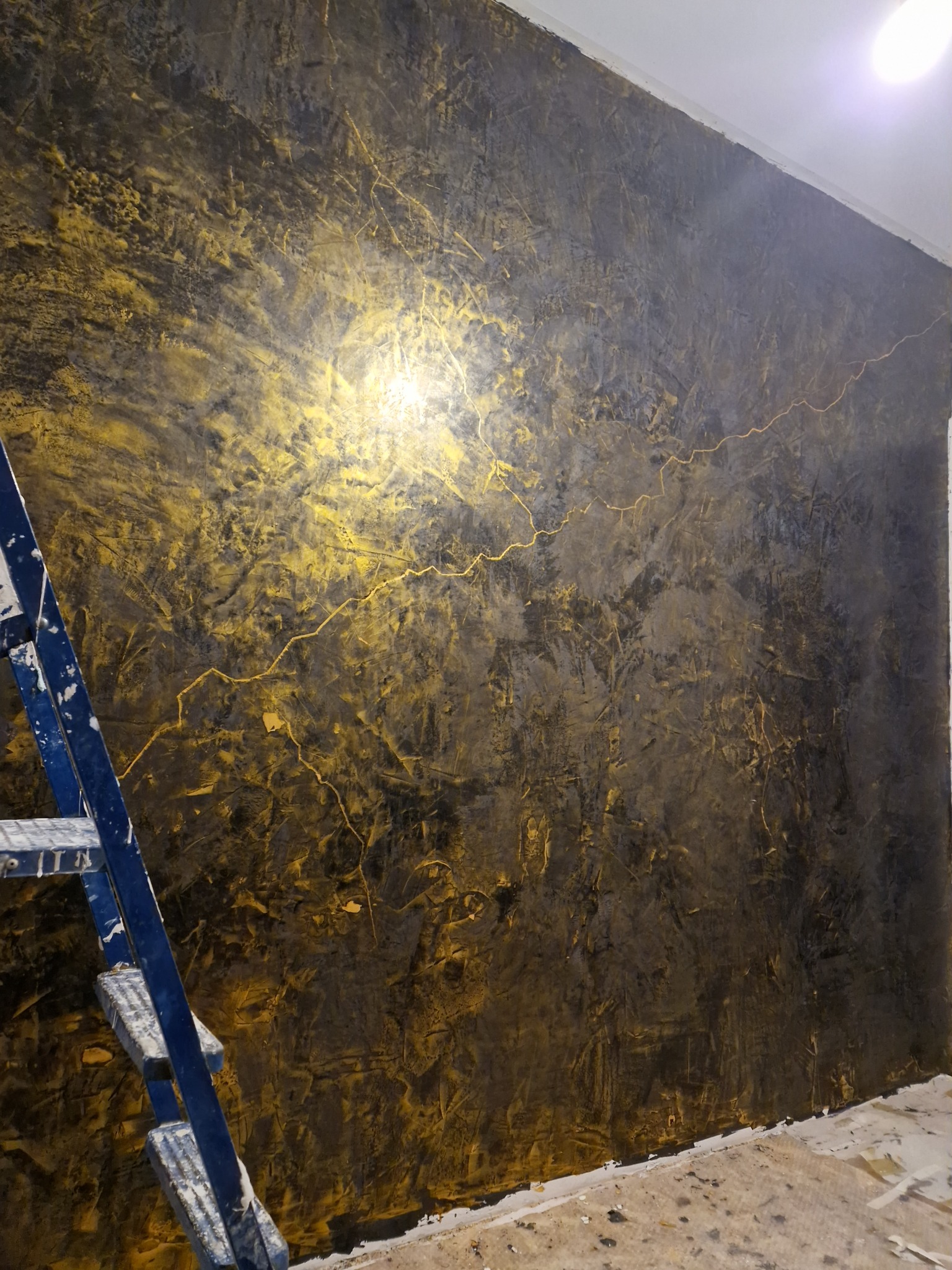 Ściana z dekoracyjną fakturą w odcieniach złota i szarości, z widoczną drabiną malarską i śladami prac wykończeniowych na podłodze.