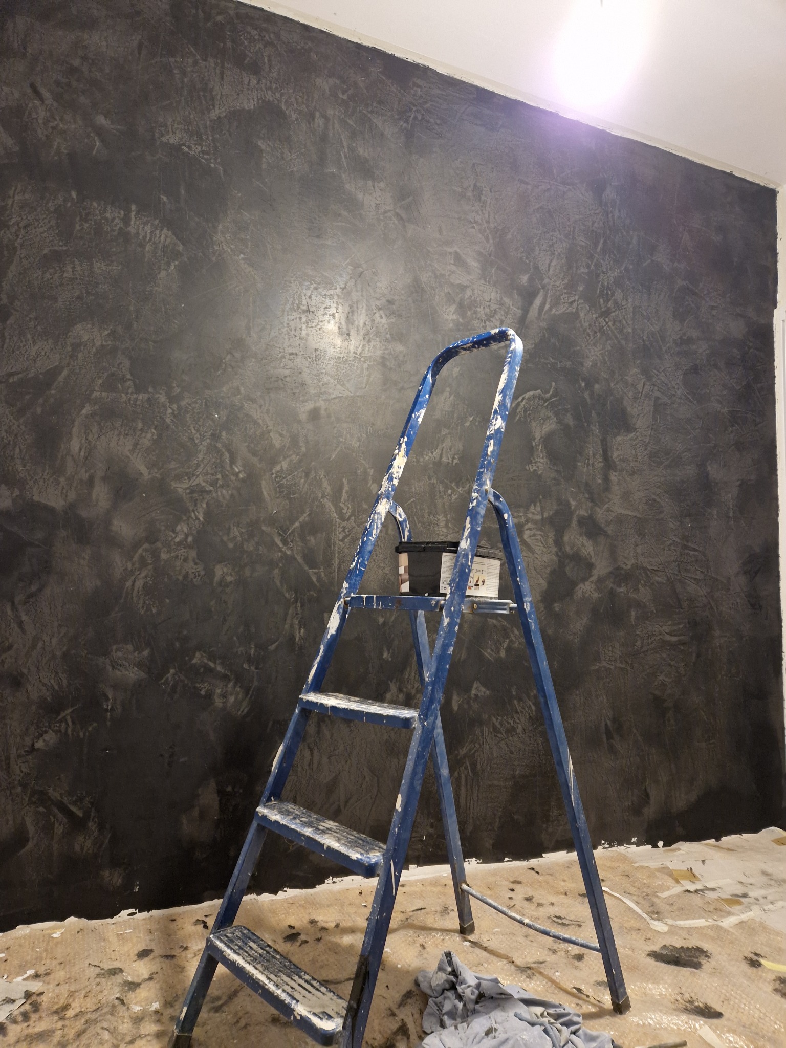 Ściana z dekoracyjną, ciemną fakturą, widoczna drabina malarska z puszką farby, fragment zabezpieczonej podłogi.