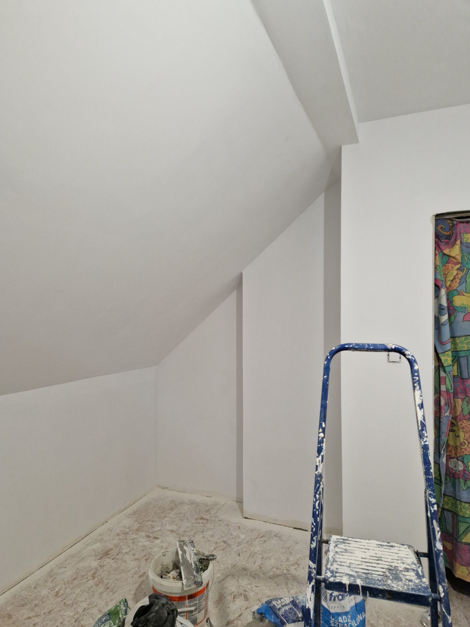 Poddasze po malowaniu na biało, widoczna niebieska drabina z resztkami farby, wiadro z narzędziami malarskimi i zasłonięte okno.