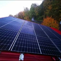 Instalacja paneli słonecznych na czerwonym dachu, widok z góry, w tle drzewa i zaparkowany samochód.