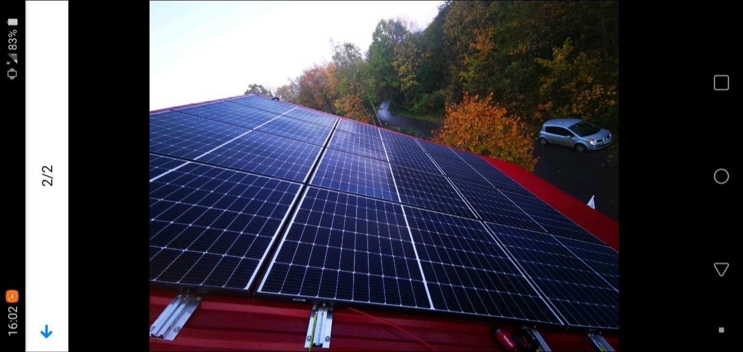 Instalacja paneli słonecznych na czerwonym dachu, widok z góry, w tle drzewa i zaparkowany samochód.