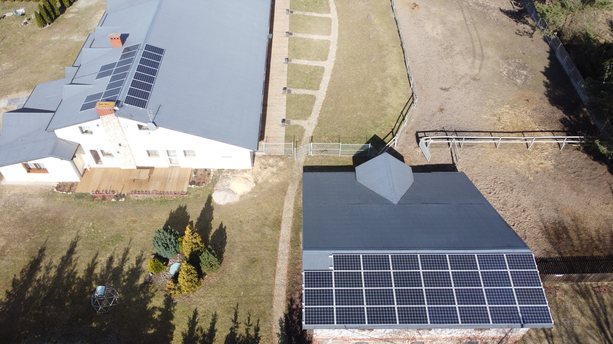 Widok z lotu ptaka na dwa budynki z panelami słonecznymi na dachach; jeden budynek biały z drewnianym tarasem, drugi ceglany z szarą dachówką. Obok budynków teren zielony i piaszczysta ścieżka.