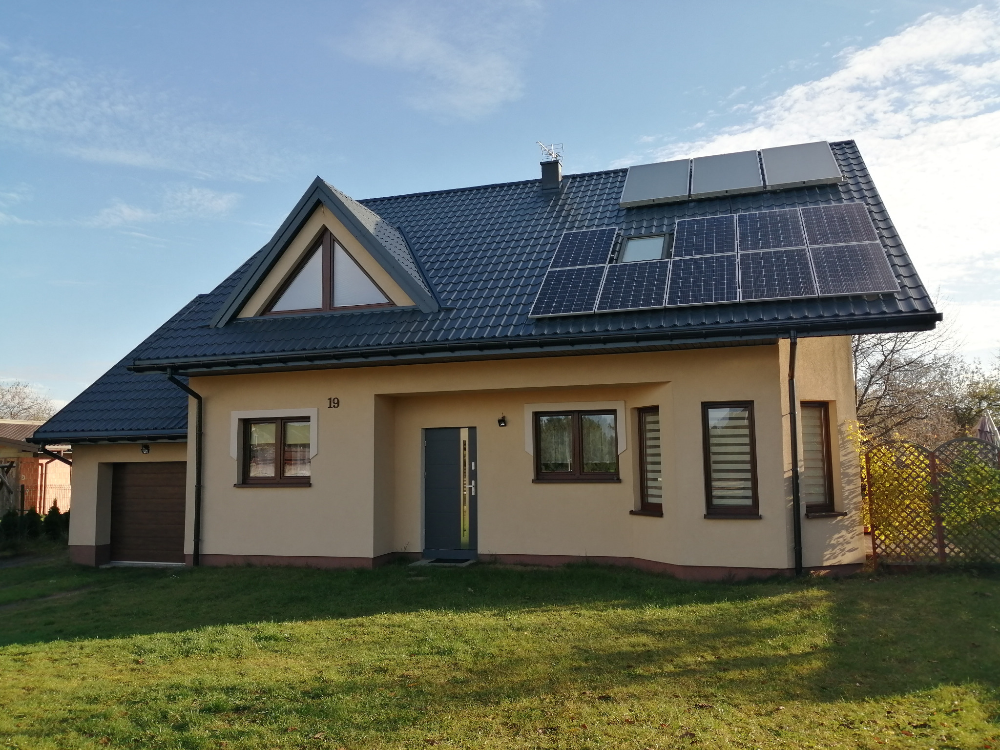 Dom jednorodzinny z panelami słonecznymi i kolektorami słonecznymi na ciemnej, skośnej połaci dachu, widok z trawnika w słoneczny dzień.