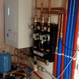 Kocioł gazowy Vaillant, zasobnik ciepłej wody, czarna skrzynka z manometrami i pompami obiegowymi, oraz instalacja z rur miedzianych i niebiesko-czerwonych rur PEX.