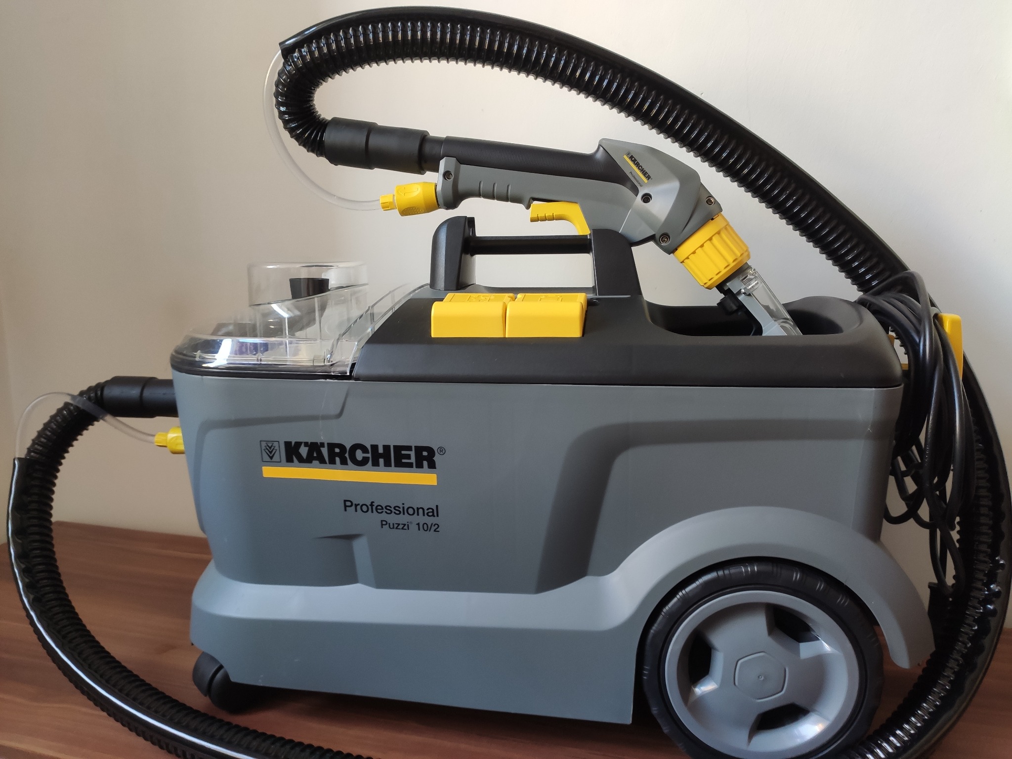 Profesjonalny odkurzacz piorący Kärcher Puzzi 10/2 z lancą i przezroczystym zbiornikiem na wodę, widoczny na drewnianej powierzchni.