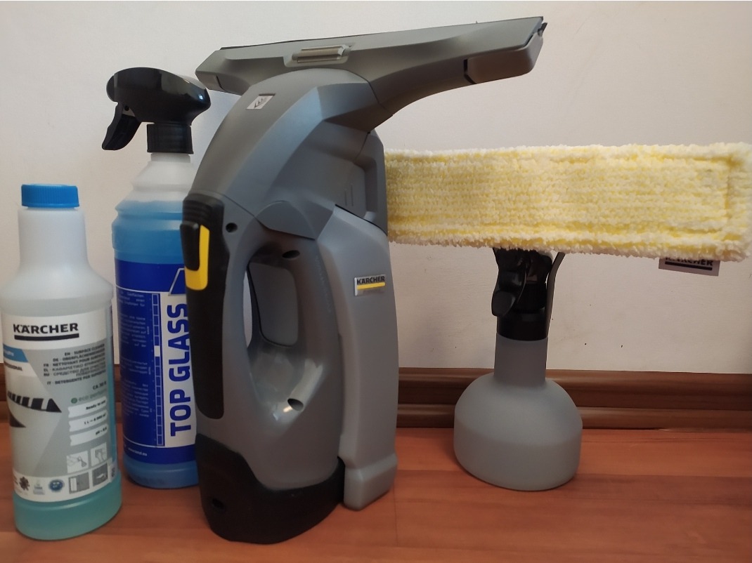 Zestaw do czyszczenia okien marki Kärcher: elektryczna myjka do okien z żółtą nakładką z mikrofibry, butelka płynu do mycia szyb Kärcher i butelka płynu Top Glass, ustawione na drewnianej podłodze.